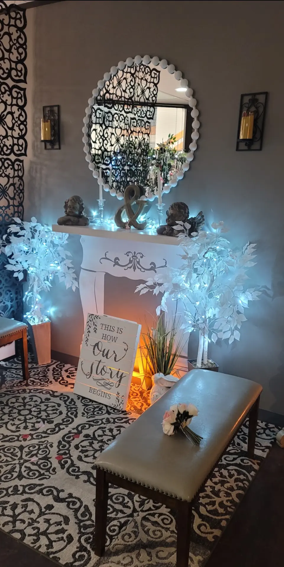 The Chapel reception fireplace setting with a candlelit mantel, ornate round mirror, glowing white flowering trees, and a 'This Is How Our Story Begins' sign.